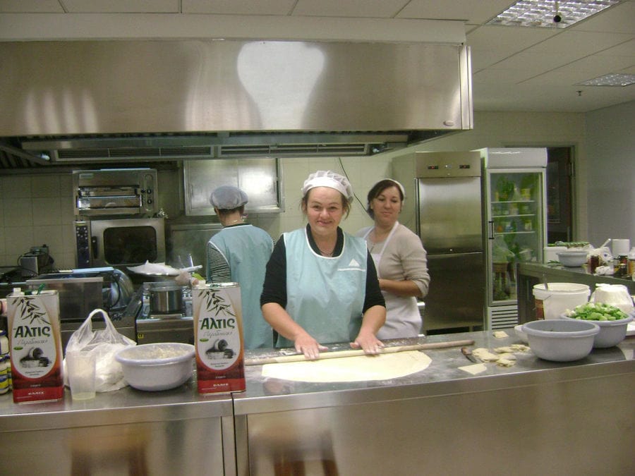 woman using wood dough paddles to laying filo dough and smiling happily at the camera